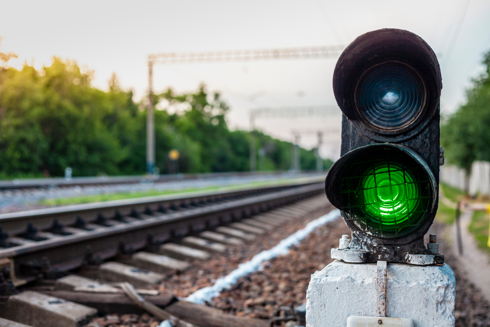 Grünes Eisenbahnsignal an einem Bahngleis in Deutschland mit unscharfem Hintergrund aus Bäumen und Schienen.